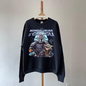 Może przedstawiać: Czarna bluza z nadrukiem The Mandalorian i Grogu. Projekt zawiera tekst "THE MANDALORIAN & GROGU" w kolorze białym i niebieskim. Obraz przedstawia postacie w ich kultowych pozach, z tłem o tematyce kosmicznej.