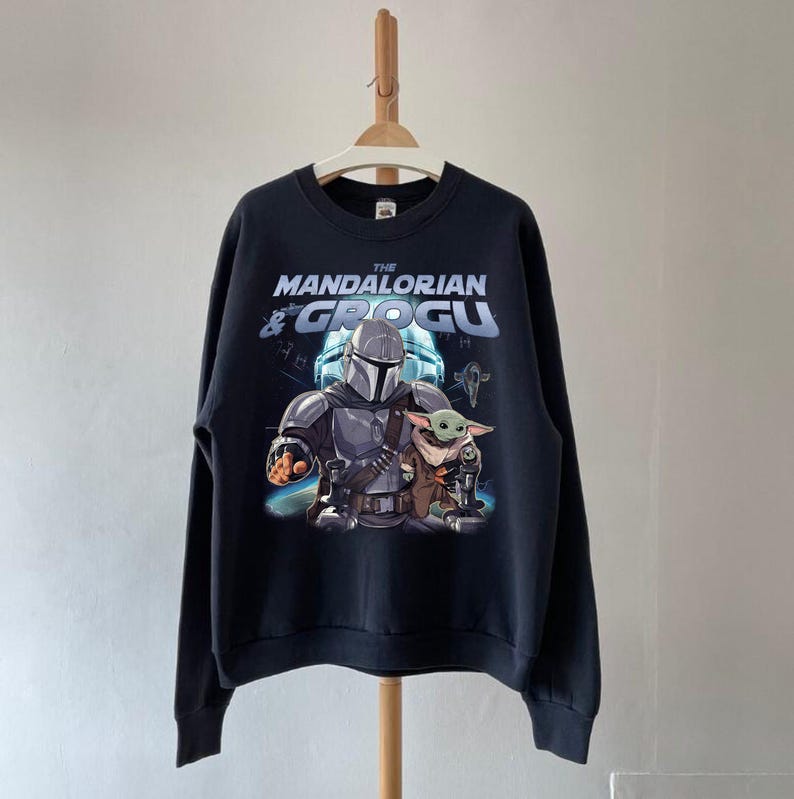 Może przedstawiać: Czarna bluza z nadrukiem The Mandalorian i Grogu. Projekt zawiera tekst "THE MANDALORIAN & GROGU" w kolorze białym i niebieskim. Obraz przedstawia postacie w ich kultowych pozach, z tłem o tematyce kosmicznej.