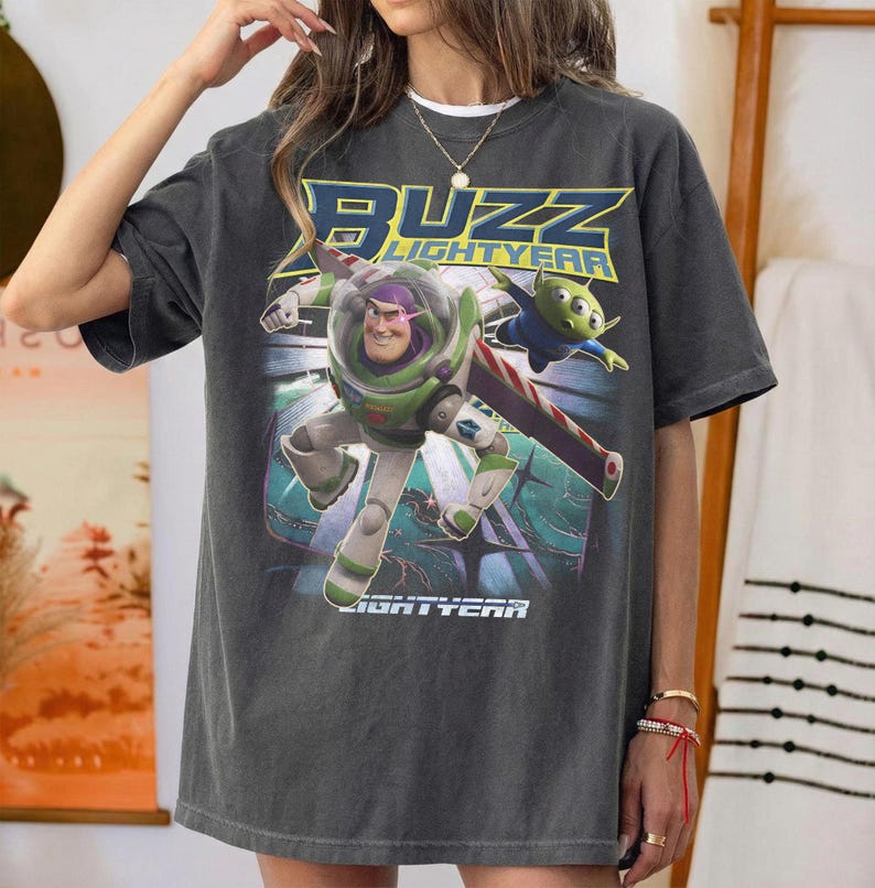 Op de afbeelding: Donkergrijs T-shirt met een afbeelding van Buzz Lightyear en een alien uit de film Lightyear. Het ontwerp bevat de tekst "BUZZ LIGHTYEAR" in geel en blauw. Het shirt heeft een relaxte, oversized pasvorm.