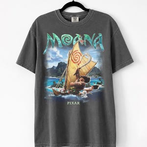 Pu&ograve; includere: T-shirt grigio scuro con una grafica del film "Moana". Il design include la parola "Moana" in turchese, una barca con una vela gialla e il logo Pixar in basso. La maglietta &egrave; appesa a una gruccia nera.