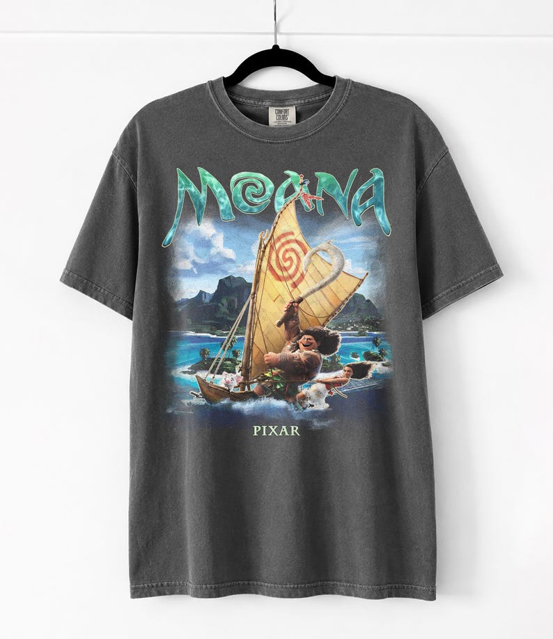 Pu&ograve; includere: T-shirt grigio scuro con una grafica del film "Moana". Il design include la parola "Moana" in turchese, una barca con una vela gialla e il logo Pixar in basso. La maglietta &egrave; appesa a una gruccia nera.