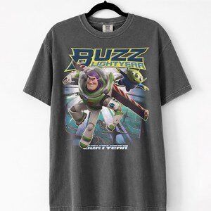 Op de afbeelding: Donkergrijs T-shirt met Buzz Lightyear uit Toy Story. De afbeelding toont Buzz in zijn ruimtepak met de woorden "Buzz Lightyear" in geel en blauw. Een groene alien is ook afgebeeld.