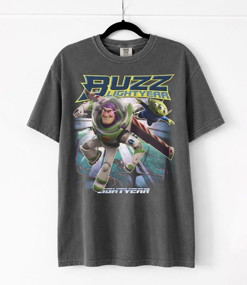 Op de afbeelding: Donkergrijs T-shirt met Buzz Lightyear uit Toy Story. De afbeelding toont Buzz in zijn ruimtepak met de woorden "Buzz Lightyear" in geel en blauw. Een groene alien is ook afgebeeld.
