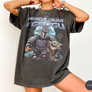 Może przedstawiać: Ciemnoszara koszulka z grafiką przedstawiającą The Mandalorian i Grogu. Projekt zawiera tekst "THE MANDALORIAN & GROGU" nad postaciami. Obrazek przedstawia Mandalorianina w zbroi i Grogu. Koszulka ma wygląd vintage.