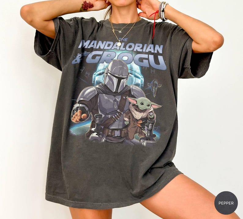 Może przedstawiać: Ciemnoszara koszulka z grafiką przedstawiającą The Mandalorian i Grogu. Projekt zawiera tekst "THE MANDALORIAN & GROGU" nad postaciami. Obrazek przedstawia Mandalorianina w zbroi i Grogu. Koszulka ma wygląd vintage.