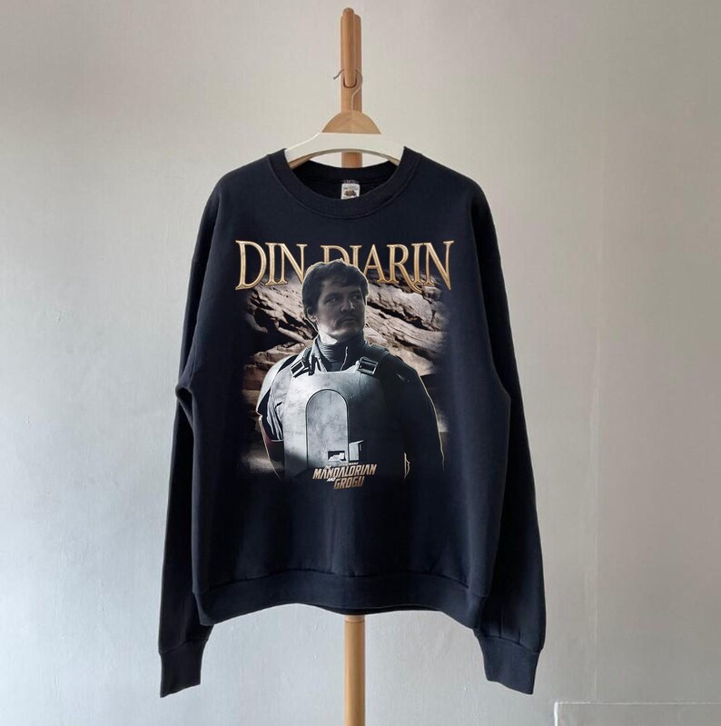 May include: Black sweatshirt featuring a graphic of Din Djarin from The Mandalorian. The text "DIN DJARIN" is above the image, and "MANDALORIAN GROGU" is below. The sweatshirt is hanging on a wooden hanger.