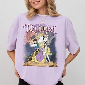 May include: A light purple t-shirt featuring a graphic of Walt Disney's Rapunzel, Flynn Rider, and Maximus. The print includes the text "Walt Disney's Rapunzel" at the top and "And at last I see the light and it's like the frog has lifted" on a banner.