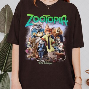 Puede incluir: Una camiseta marr&oacute;n oscuro con un gr&aacute;fico colorido de la pel&iacute;cula animada "Zootopia". El dise&ntilde;o incluye el t&iacute;tulo de la pel&iacute;cula en verde y azul, junto con varias ilustraciones de personajes. Tambi&eacute;n es visible el logotipo de Walt Disney.