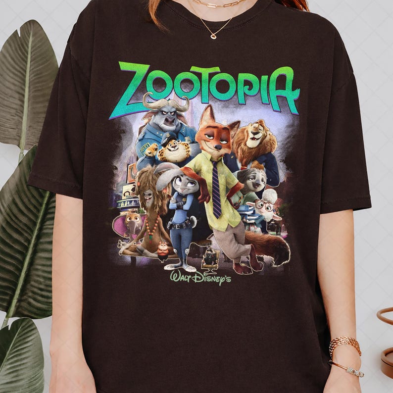 Puede incluir: Una camiseta marr&oacute;n oscuro con un gr&aacute;fico colorido de la pel&iacute;cula animada "Zootopia". El dise&ntilde;o incluye el t&iacute;tulo de la pel&iacute;cula en verde y azul, junto con varias ilustraciones de personajes. Tambi&eacute;n es visible el logotipo de Walt Disney.