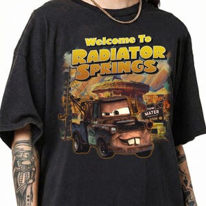 Puede incluir: Camiseta gris oscuro con la frase "Welcome To Radiator Springs" en amarillo y naranja. El gr&aacute;fico incluye una ilustraci&oacute;n de Mate de la pel&iacute;cula Cars. La camiseta es de cuello redondo.