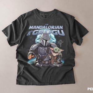 Może przedstawiać: Ciemnoszara koszulka z grafiką The Mandalorian i Grogu z serii Star Wars. Projekt zawiera tekst "The Mandalorian & Grogu" w kolorze niebieskim i białym, z ilustracjami postaci.