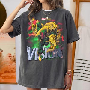 Puede incluir: Camiseta gris oscuro con un gr&aacute;fico del personaje de Marvel, Vision. El dise&ntilde;o incluye a Vision en verde y rojo, con detalles amarillos, y el texto "THE VISION" en blanco. El logotipo de Marvel Studios est&aacute; en la parte superior.