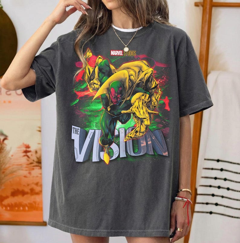 Puede incluir: Camiseta gris oscuro con un gr&aacute;fico del personaje de Marvel, Vision. El dise&ntilde;o incluye a Vision en verde y rojo, con detalles amarillos, y el texto "THE VISION" en blanco. El logotipo de Marvel Studios est&aacute; en la parte superior.