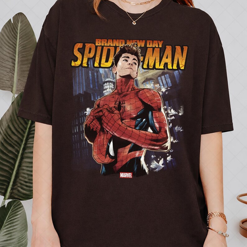 Pu&ograve; includere: T-shirt marrone scuro con grafica di Spider-Man e la scritta "BRAND NEW DAY SPIDER-MAN" in giallo e arancione. Il logo Marvel &egrave; in basso. Il design include un paesaggio urbano e uccelli bianchi.