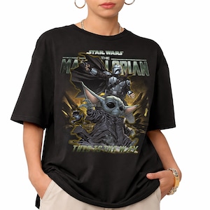 May include: Black t-shirt featuring a graphic from the Star Wars series, The Mandalorian. The design includes the Mandalorian, Grogu, and the text "This is the Way" in a yellow and green color scheme.
