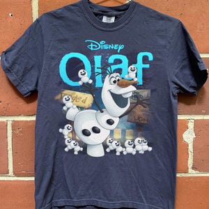 May include: Navy blue t-shirt featuring the Disney character Olaf from Frozen. The design includes Olaf, small snowmen, and the word "Olaf" in turquoise. The shirt is hanging on a metal hanger against a brick wall.