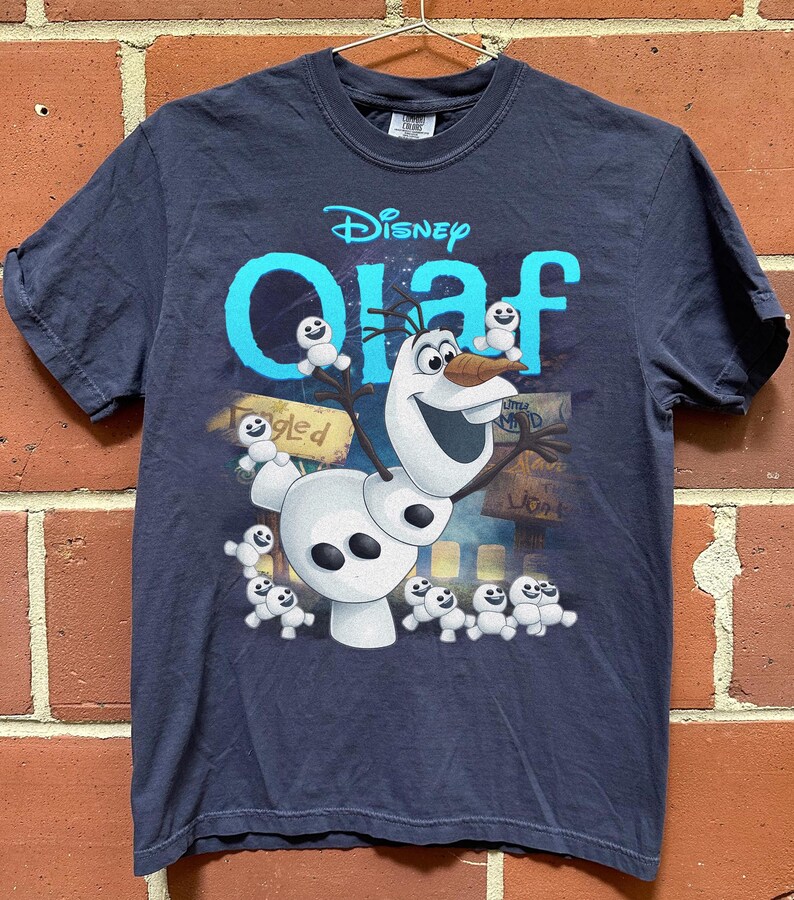 May include: Navy blue t-shirt featuring the Disney character Olaf from Frozen. The design includes Olaf, small snowmen, and the word "Olaf" in turquoise. The shirt is hanging on a metal hanger against a brick wall.