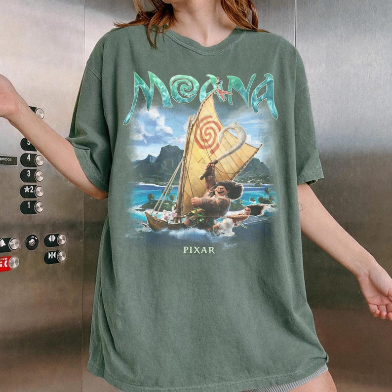 Pu&ograve; includere: T-shirt verde con la scritta "Moana" in lettere turchesi. La maglietta presenta un'illustrazione colorata di una barca che naviga sull'oceano con un personaggio del film. La parola "PIXAR" &egrave; stampata sotto.