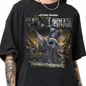 May include: Dark gray t-shirt featuring a graphic from the Star Wars series, "The Mandalorian." The design includes the Mandalorian, a character in silver armor, and Grogu, also known as Baby Yoda. The text "This is the Way" is also visible.