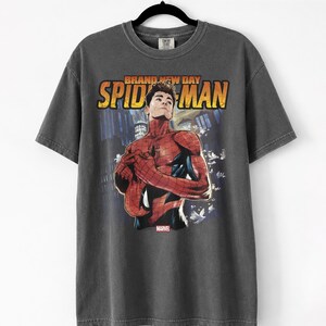 Pu&ograve; includere: T-shirt grigio scuro con una grafica di Spider-Man e la scritta "BRAND NEW DAY SPIDER-MAN" in arancione e giallo. Il logo Marvel &egrave; visibile. La maglietta &egrave; appesa a una gruccia nera.