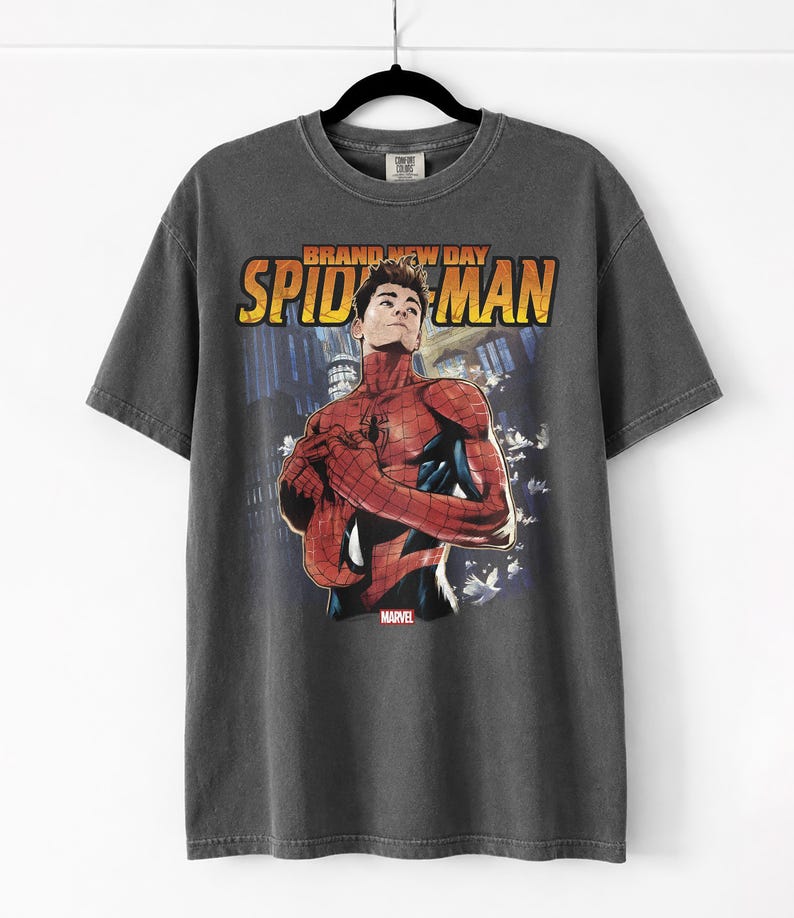 Pu&ograve; includere: T-shirt grigio scuro con una grafica di Spider-Man e la scritta "BRAND NEW DAY SPIDER-MAN" in arancione e giallo. Il logo Marvel &egrave; visibile. La maglietta &egrave; appesa a una gruccia nera.