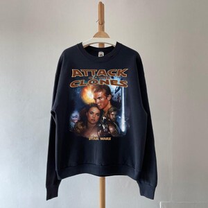 May include: Black sweatshirt with the movie poster for Star Wars: Attack of the Clones. The design includes the film's title in gold lettering, and images of the main characters. The sweatshirt is hanging on a wooden hanger.