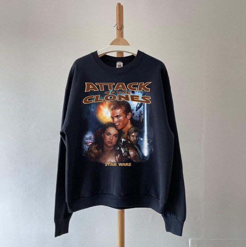 May include: Black sweatshirt with the movie poster for Star Wars: Attack of the Clones. The design includes the film's title in gold lettering, and images of the main characters. The sweatshirt is hanging on a wooden hanger.