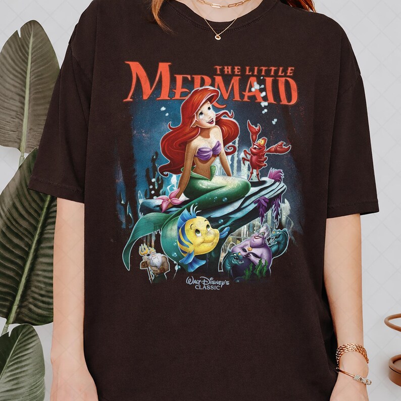 May include: A dark brown t-shirt featuring a colorful graphic from Disney's "The Little Mermaid." The design includes Ariel, Flounder, Sebastian, and other characters, with the title in red above the scene. The shirt is a classic.
