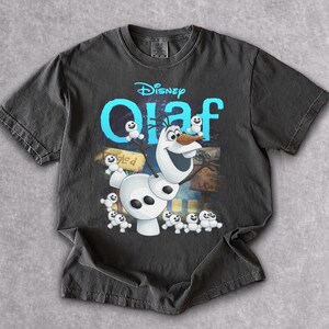 May include: Dark gray t-shirt featuring the Disney character Olaf from Frozen. The design includes Olaf, small snowmen, and the word "Olaf" in blue. The shirt is made of a soft material.