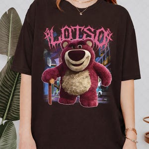 K&ouml;nnte beinhalten: Dunkelbraunes T-Shirt mit einer Grafik von Lotso, einem rosa B&auml;rencharakter, und dem Wort "LOTSO" in rosa, stilisiertem Text. Der B&auml;r h&auml;lt einen Holzstock. Der Hintergrund zeigt eine Zeichentrickszene.