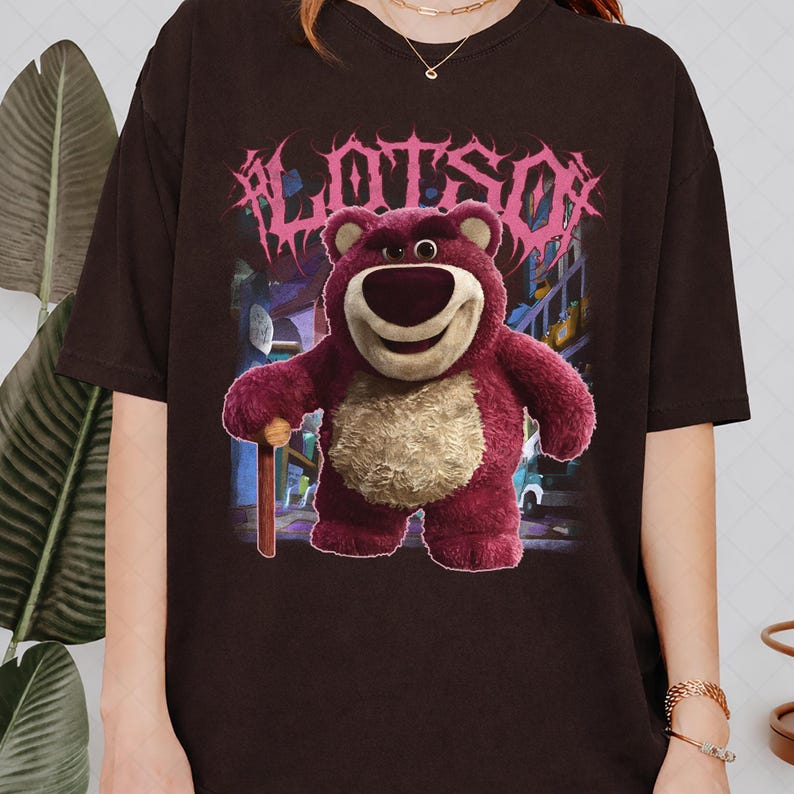 K&ouml;nnte beinhalten: Dunkelbraunes T-Shirt mit einer Grafik von Lotso, einem rosa B&auml;rencharakter, und dem Wort "LOTSO" in rosa, stilisiertem Text. Der B&auml;r h&auml;lt einen Holzstock. Der Hintergrund zeigt eine Zeichentrickszene.
