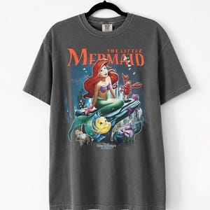 May include: Dark gray t-shirt featuring a colorful graphic of Ariel from Disney's The Little Mermaid. The design includes Ariel, Flounder, Sebastian, and other characters, with the words "The Little Mermaid" in orange. The shirt is a classic Disney design.