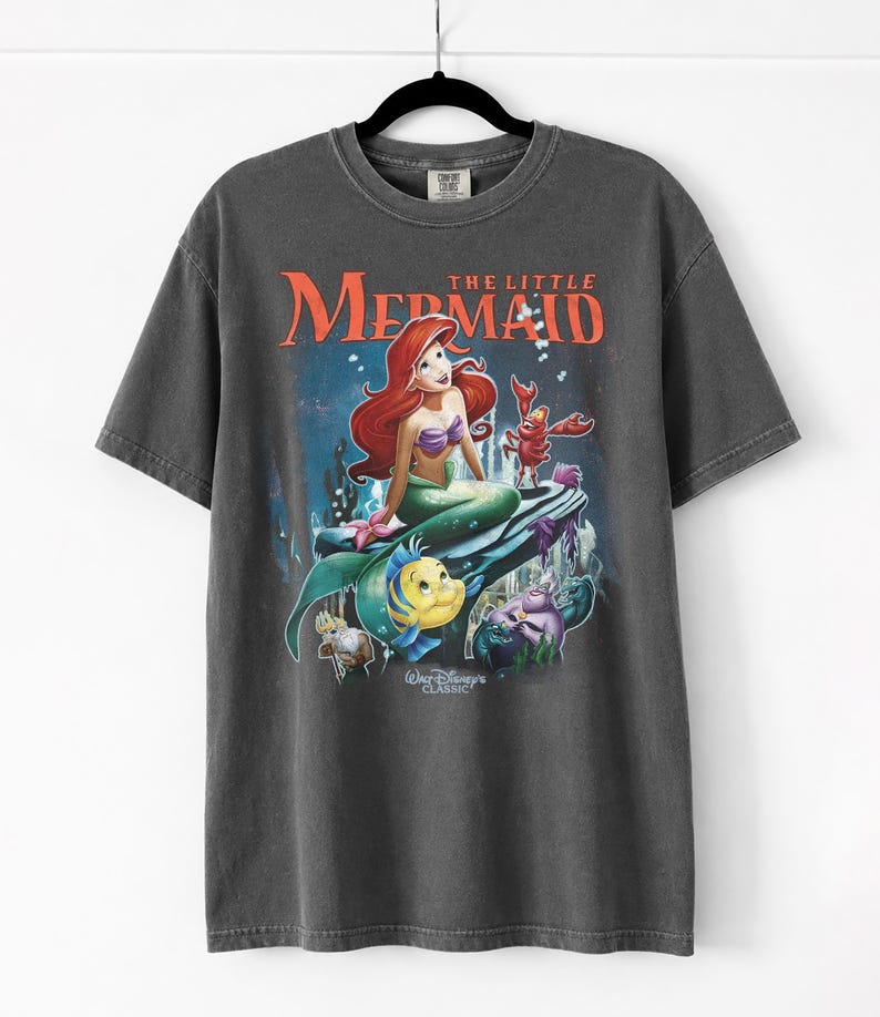 May include: Dark gray t-shirt featuring a colorful graphic of Ariel from Disney's The Little Mermaid. The design includes Ariel, Flounder, Sebastian, and other characters, with the words "The Little Mermaid" in orange. The shirt is a classic Disney design.