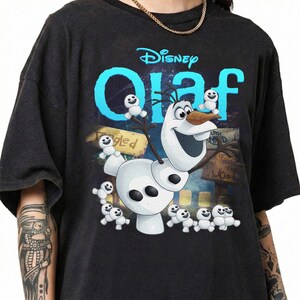 May include: Black t-shirt featuring the Disney character Olaf from Frozen. The design includes Olaf, small snowmen, and the word "Olaf" in blue, with the Disney logo above. The shirt has short sleeves.