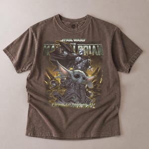 May include: Brown Star Wars The Mandalorian t-shirt featuring a graphic of The Mandalorian and Grogu. The text "STAR WARS THE MANDALORIAN" is printed across the top. The shirt is made of a soft, comfortable material.