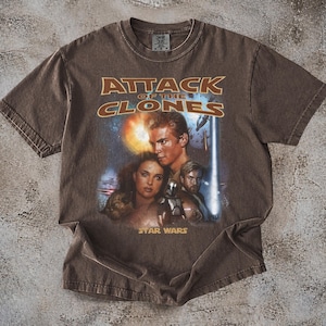 May include: Brown t-shirt with the text "ATTACK OF THE CLONES" and a graphic of Star Wars characters. The Star Wars logo is below the graphic. The shirt has a crew neck and short sleeves.