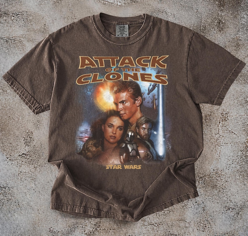May include: Brown t-shirt with the text "ATTACK OF THE CLONES" and a graphic of Star Wars characters. The Star Wars logo is below the graphic. The shirt has a crew neck and short sleeves.