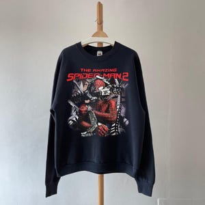 May include: Black sweatshirt featuring a graphic from "The Amazing Spider-Man 2". The design includes Spider-Man in a red and black suit, with the movie title in red and white lettering. The sweatshirt is hanging on a wooden hanger.