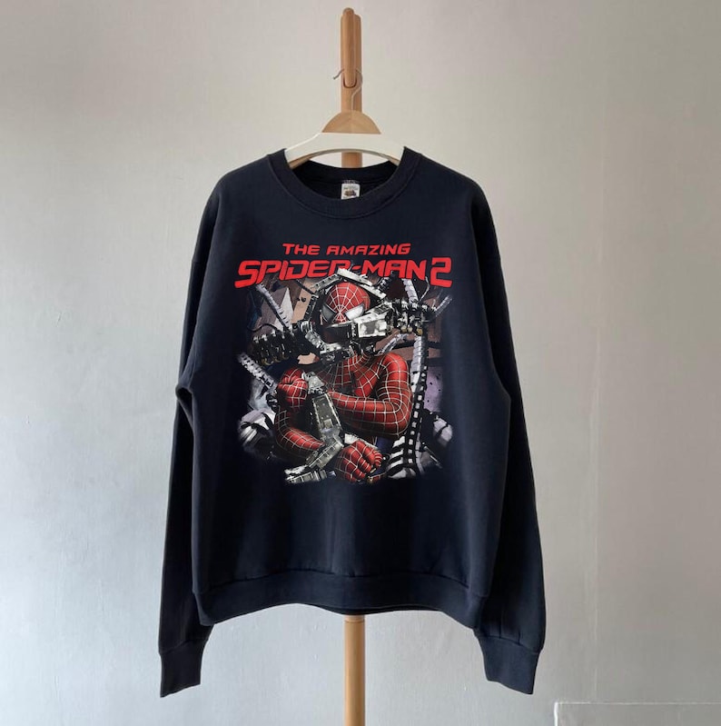May include: Black sweatshirt featuring a graphic from "The Amazing Spider-Man 2". The design includes Spider-Man in a red and black suit, with the movie title in red and white lettering. The sweatshirt is hanging on a wooden hanger.