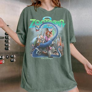 May include: Green t-shirt with a graphic from the film "Zootropolis." The design includes the film's title, a blue snake, and characters Nick Wilde and Judy Hopps. The Walt Disney's logo is at the bottom.