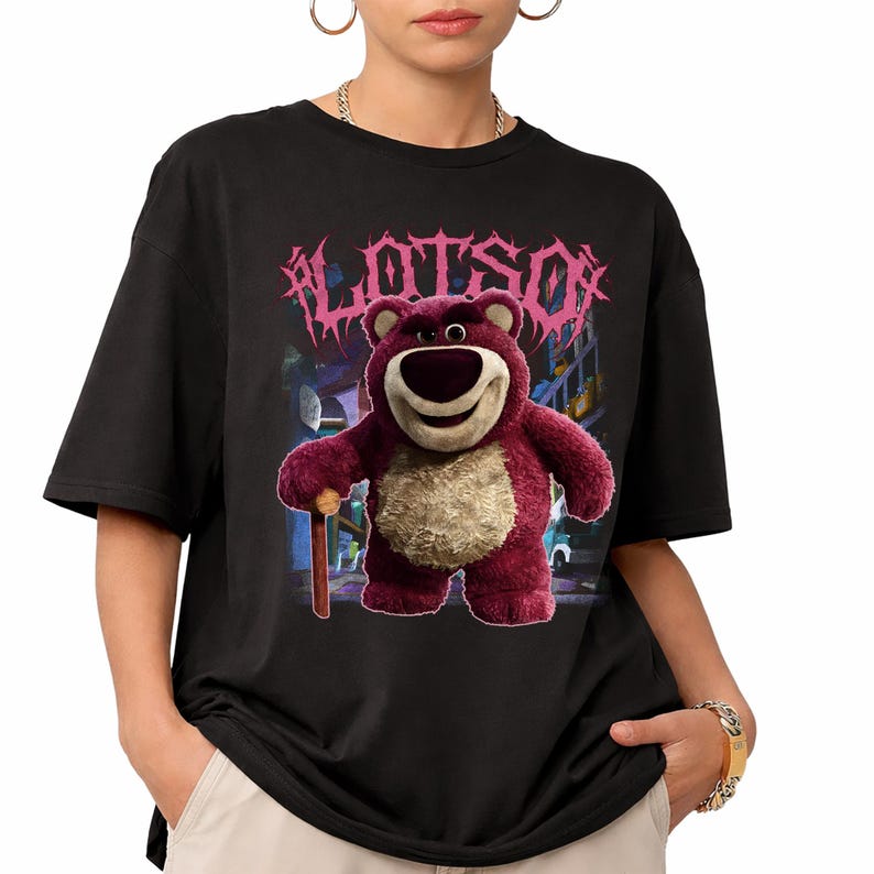 K&ouml;nnte beinhalten: Schwarzes T-Shirt mit einem Cartoon-Bild von Lotso, einem rosa B&auml;rencharakter, und dem Wort "LOTSO" in rosa, stilisiertem Text &uuml;ber dem B&auml;ren. Der B&auml;r h&auml;lt einen Holzstock. Der Hintergrund ist eine verschwommene Stadtszene.
