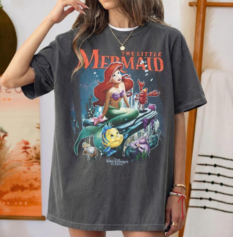May include: Dark gray t-shirt featuring a graphic of Ariel from Disney's "The Little Mermaid." The design includes Ariel, Flounder, Sebastian, and other characters. The text "The Little Mermaid" is in orange. "Walt Disney's Classic" is also printed on the shirt.