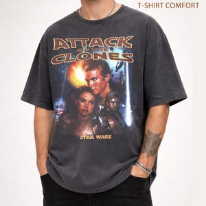 May include: Charcoal grey t-shirt with a movie poster design for "Attack of the Clones" from the Star Wars franchise. The graphic features characters and the film's title in a distressed style. The text "Pepper T-Shirt Comfort" is printed at the top.