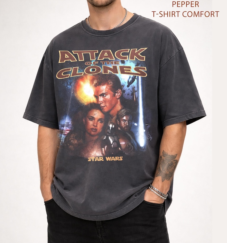 May include: Charcoal grey t-shirt with a movie poster design for "Attack of the Clones" from the Star Wars franchise. The graphic features characters and the film's title in a distressed style. The text "Pepper T-Shirt Comfort" is printed at the top.