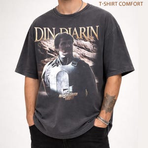 May include: Dark gray t-shirt with a distressed graphic of Din Djarin from The Mandalorian. The text "DIN DJARIN" is in gold lettering above the image. The shirt also features the text "MANDALORIAN GROGU" below the image. The shirt is labeled "PEPPER T-SHIRT COMFORT".