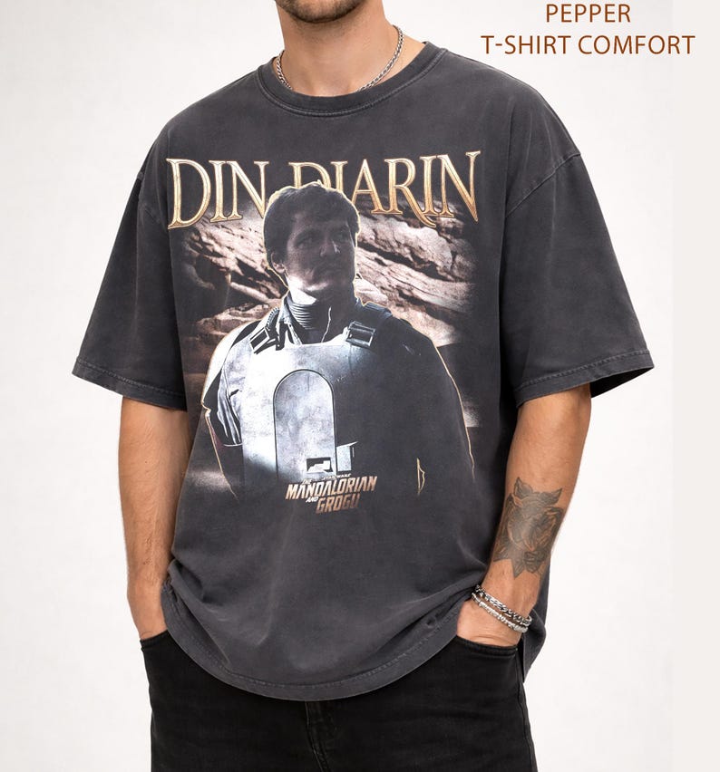 May include: Dark gray t-shirt with a distressed graphic of Din Djarin from The Mandalorian. The text "DIN DJARIN" is in gold lettering above the image. The shirt also features the text "MANDALORIAN GROGU" below the image. The shirt is labeled "PEPPER T-SHIRT COMFORT".