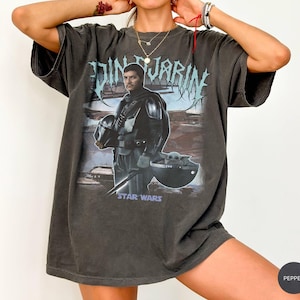 May include: Dark gray t-shirt featuring a graphic of Din Djarin from Star Wars, holding his helmet, with Grogu in a floating pod. The text "Din Djarin" is above the image in a stylized font, and "Star Wars" is below.