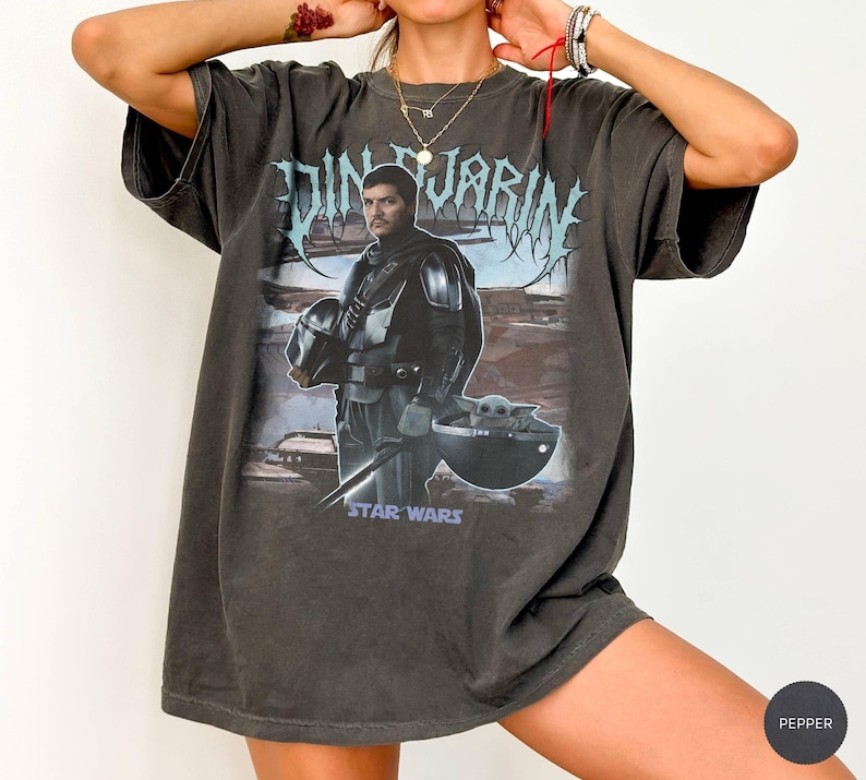 May include: Dark gray t-shirt featuring a graphic of Din Djarin from Star Wars, holding his helmet, with Grogu in a floating pod. The text "Din Djarin" is above the image in a stylized font, and "Star Wars" is below.