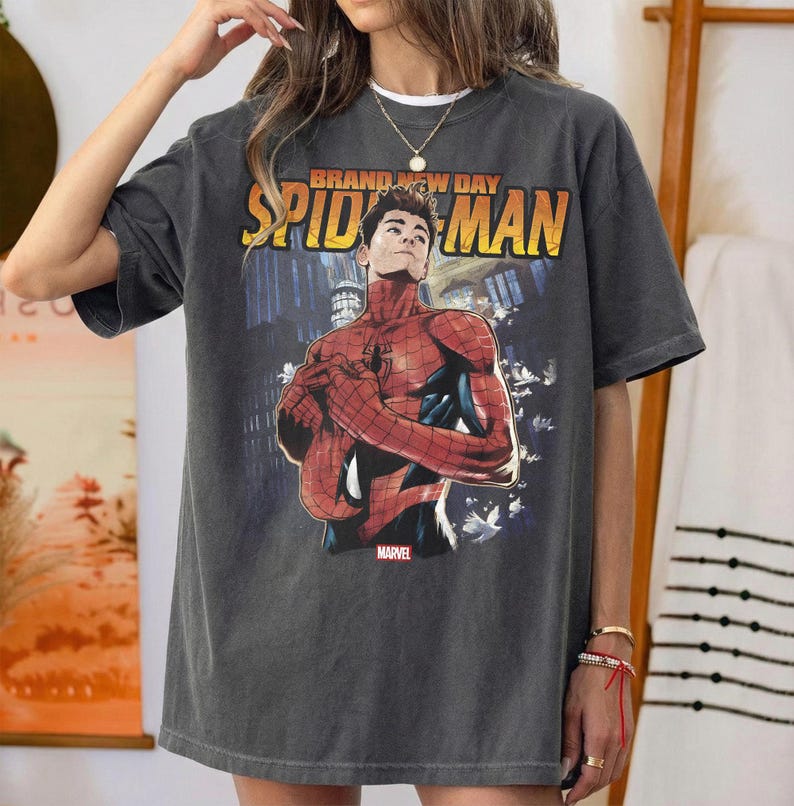 Pu&ograve; includere: T-shirt grigio scuro con grafica di Spider-Man e la scritta "BRAND NEW DAY SPIDER-MAN" in giallo e arancione. Il logo Marvel &egrave; in basso. La maglietta ha un aspetto vintage.