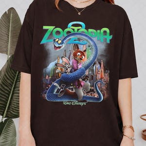 May include: A dark brown t-shirt with a vibrant graphic from the film Zootopia. The design showcases the film's title, a blue snake, and the characters Judy Hopps and Nick Wilde. The Walt Disney's logo is also visible.